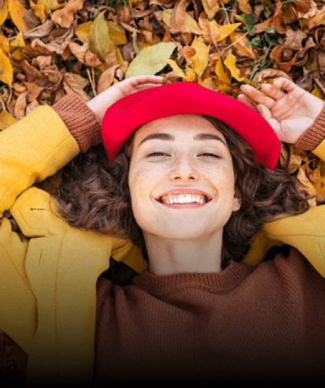 Smiling woman in red hat lying on autumn leaves wearing yellow and brown clothing.
