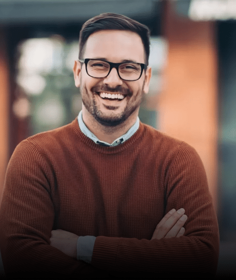 Smiling male dental professional wearing glasses and brown sweater in front of blurred office building.