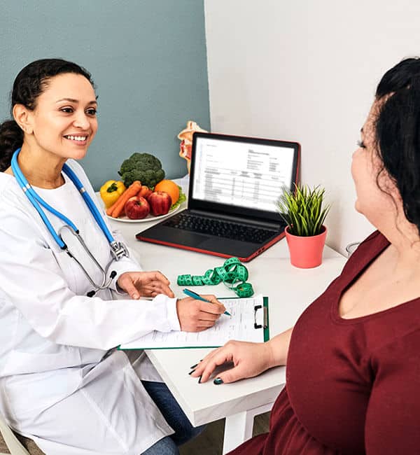 Female dentist in white coat consults with patient in clinical office setting with laptop.