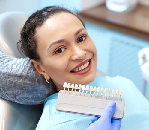 Woman smiling in dental chair holding tooth shade guide at dental office.