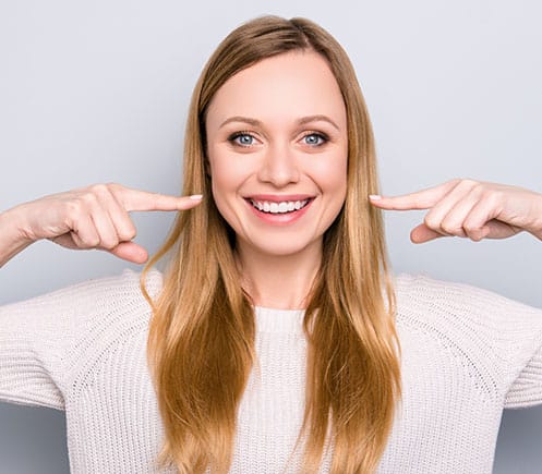 Smiling woman pointing to her teeth against neutral background for dental practice website.