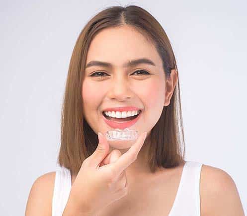 Young woman smiling brightly while touching her teeth against white background.