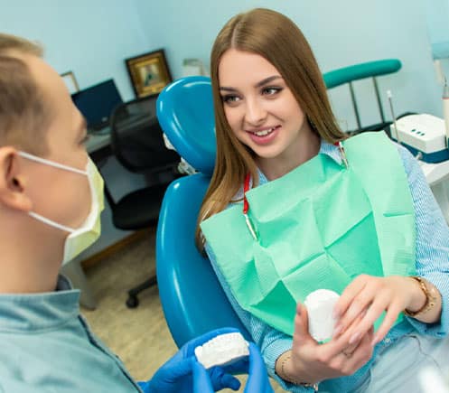 Female dental professional in green scrubs holding model teeth, seated in dental office chair.