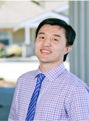 Young male professional in purple checkered shirt and tie smiling in modern dental office setting.